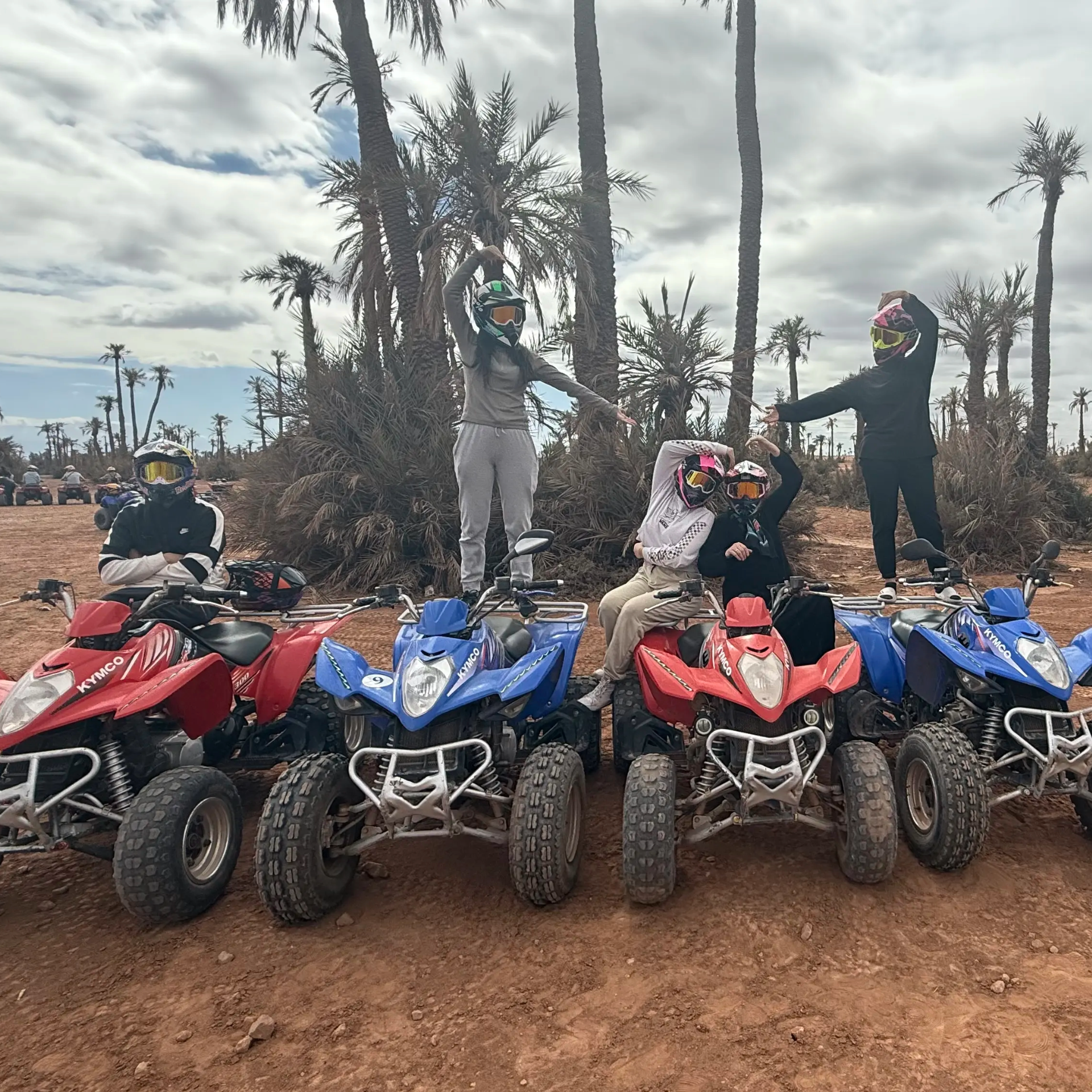 Groupe de quatre personnes avec quatre quads, deux rouges et deux bleus, devant les palmiers de la palmeraie de Marrakech sous un ciel bleu nuageux