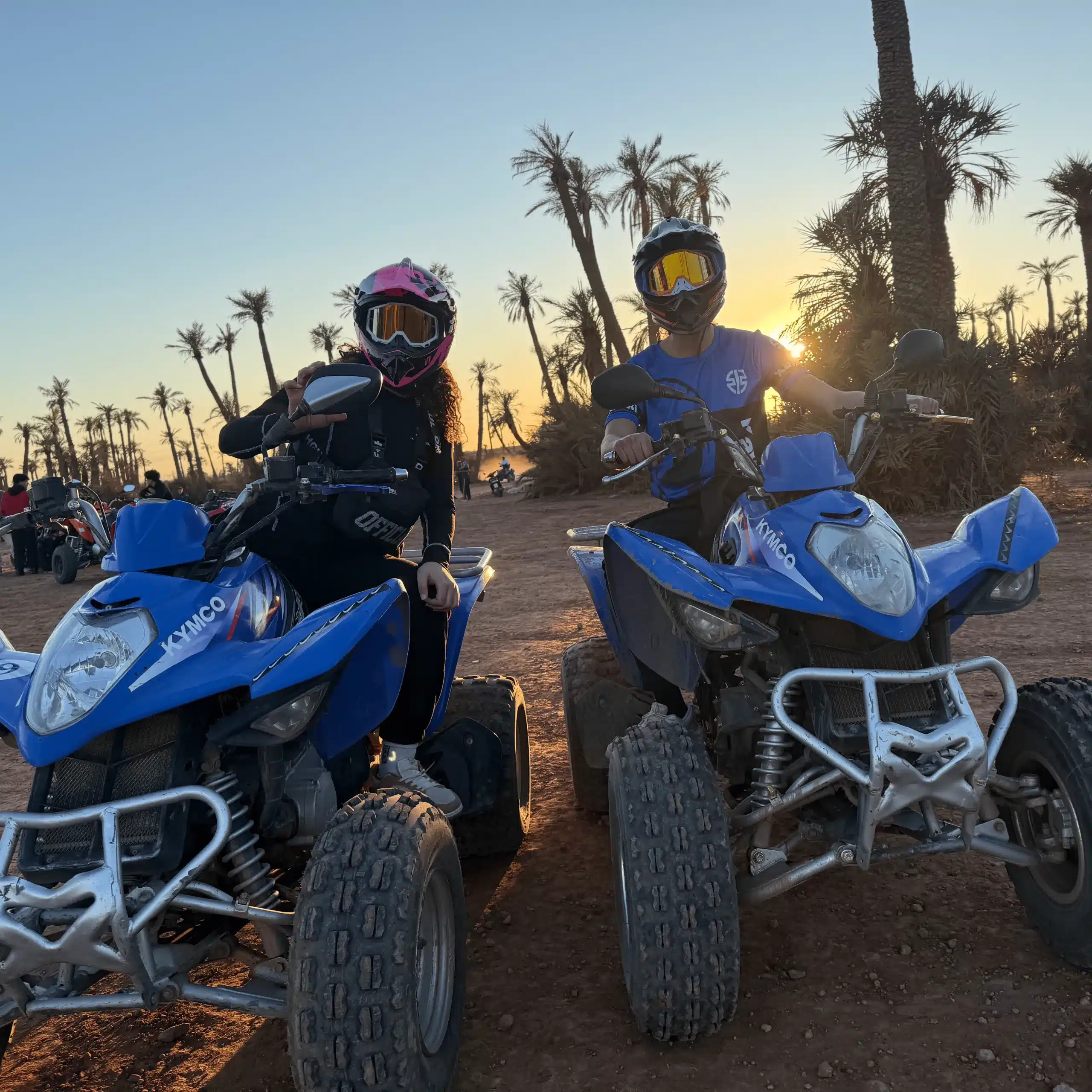 Couple sur quads Kymco Maxxer 300 bleus, casqués, dans la palmeraie de Marrakech au coucher du soleil