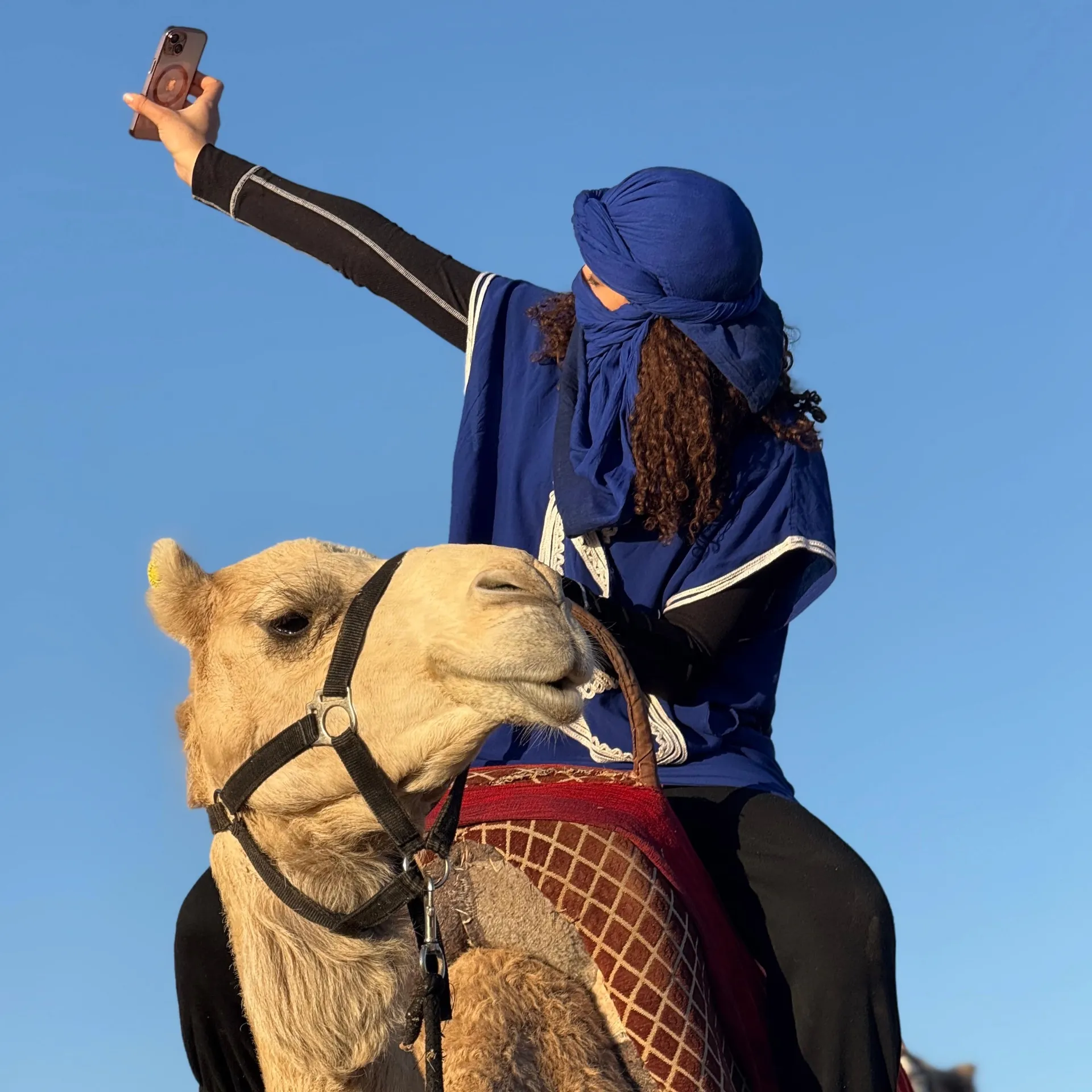 Femme sur un dromadaire à la Palmeraie de Marrakech, portant une tenue bleue avec un chèche traditionnel, prenant un selfie