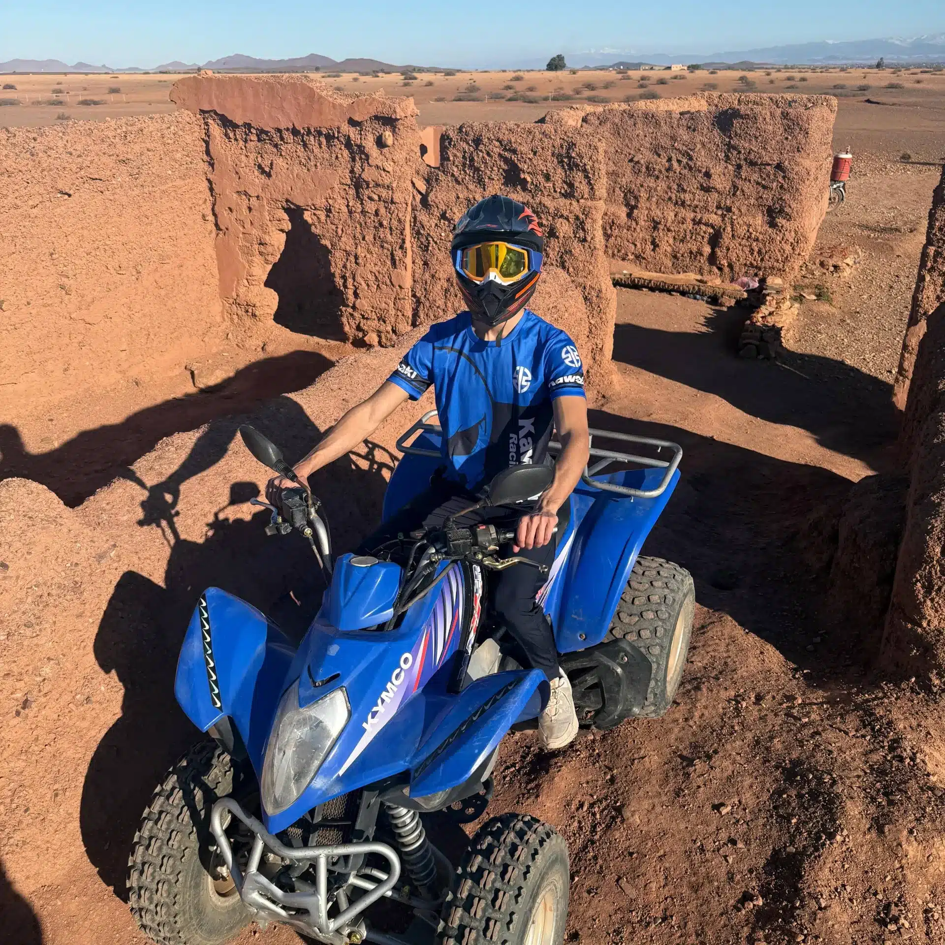 Pilote casqué sur quad Kymco Maxxer 300 bleu dans la palmeraie de Marrakech, murs sable rappelant un château en arrière-plan